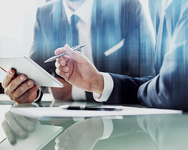 Two people in business suits sit at a desk, discussing Oil Plant Engineering Services India, as one holds a digital tablet and the other points at it with a pen at Sungates Technologies Pvt Ltd.